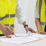 Two construction professionals wearing high-visibility vests reviewing building regulation documents on a table.