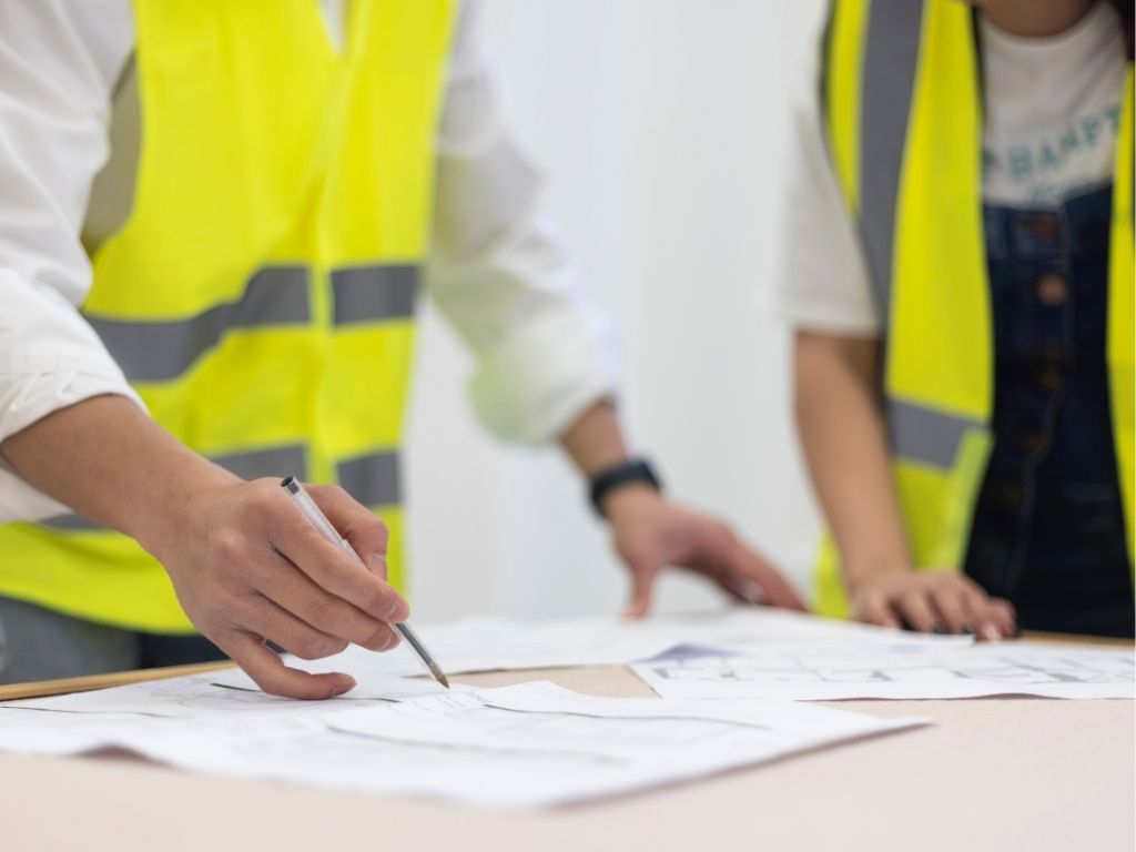 Two construction professionals wearing high-visibility vests reviewing building regulation documents on a table.